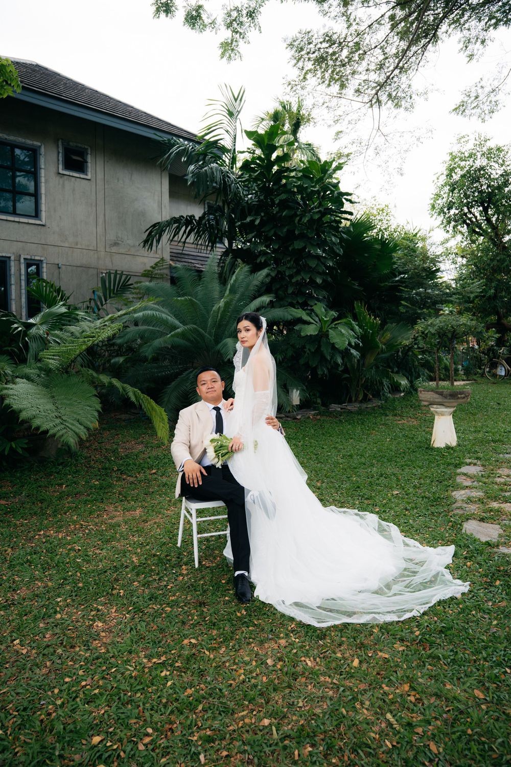 Bride in white gown and groom in suit pose in lush garden setting outside elegant venue.