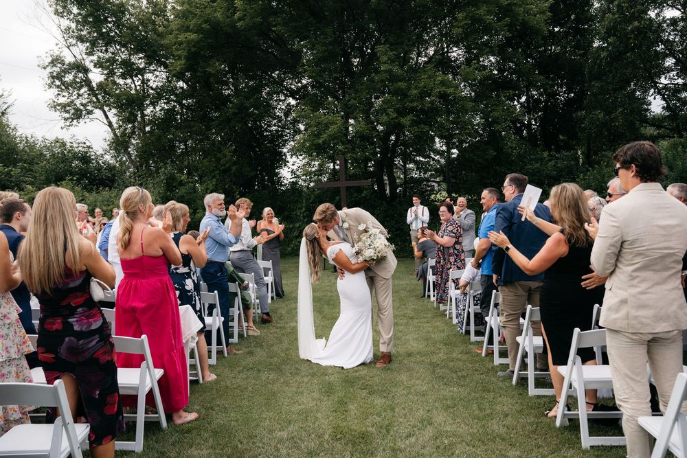 Outdoor wedding ceremony with bride and groom kissing in aisle, guests applauding on white chairs, lush green trees backdrop.