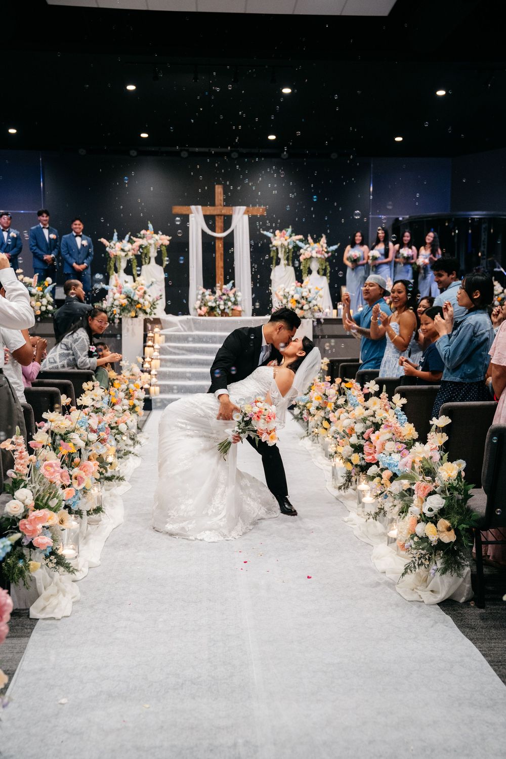 Bride and groom sharing romantic dip kiss at church altar exit lined with colorful floral arrangements.