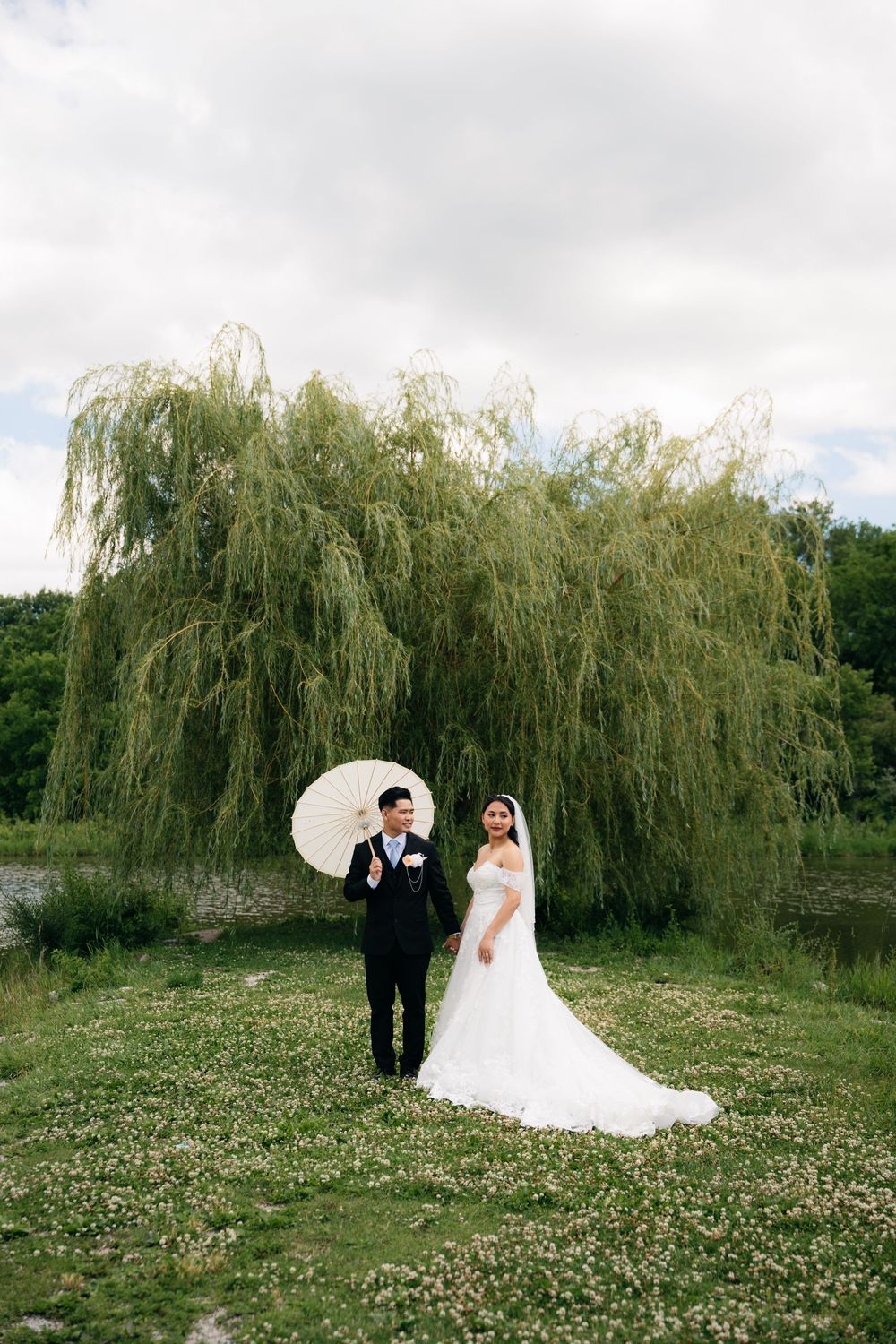 Asian bride and groom holding white parasol standing in wildflower field beneath weeping willow tree.
