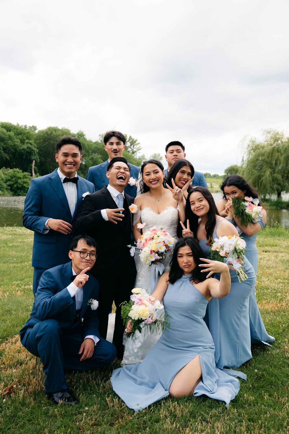 Wedding party in blue suits and dresses posing playfully with bride and groom near lakeside.