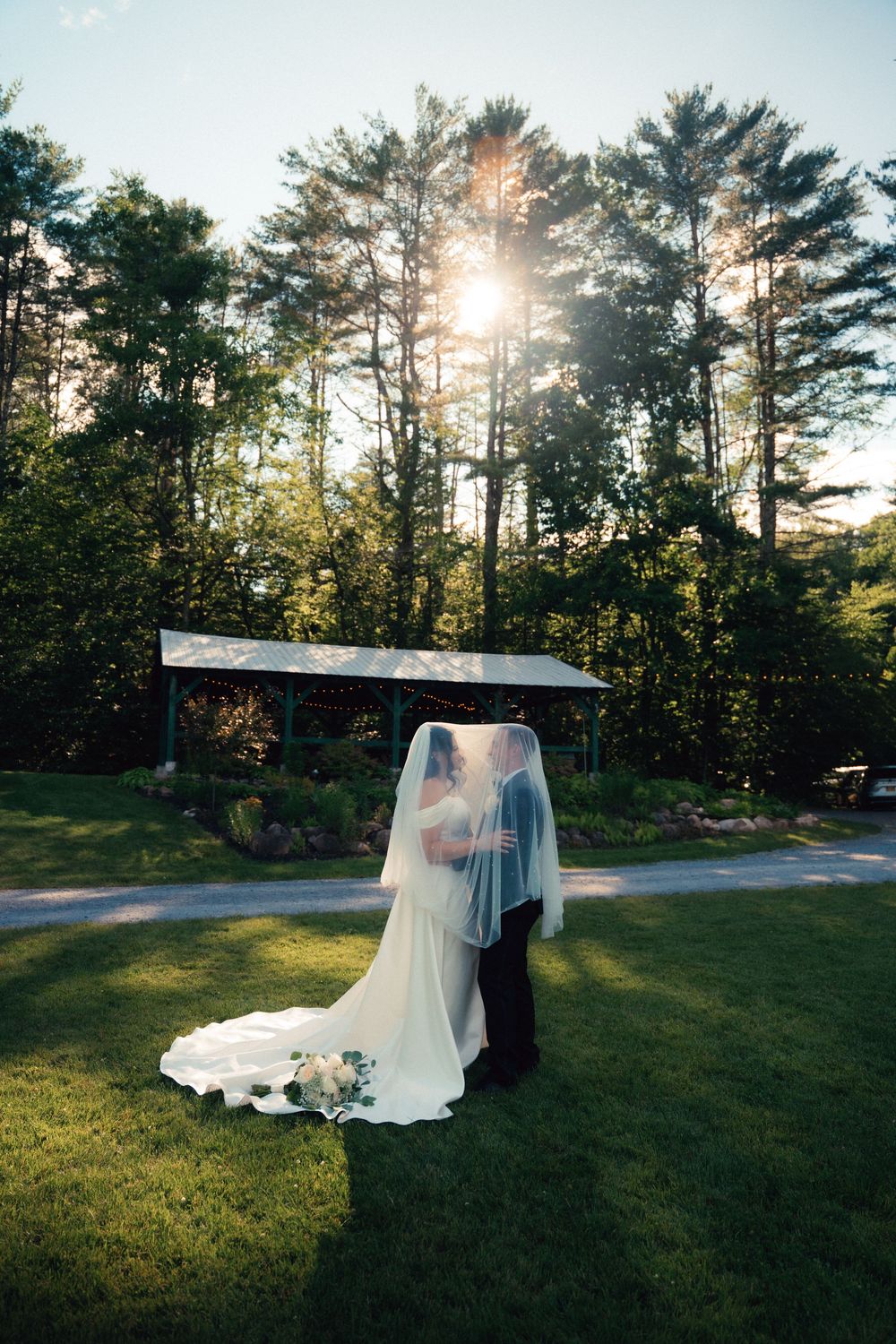 Bride and groom share a kiss under a flowing veil on a lush green lawn with sunlight filtering through tall trees.