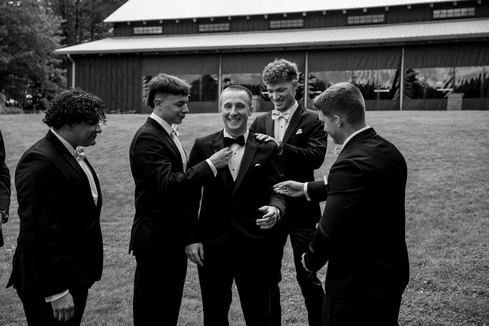 Groomsmen in black tuxedos laugh and celebrate together outdoors near a barn in black and white photo.