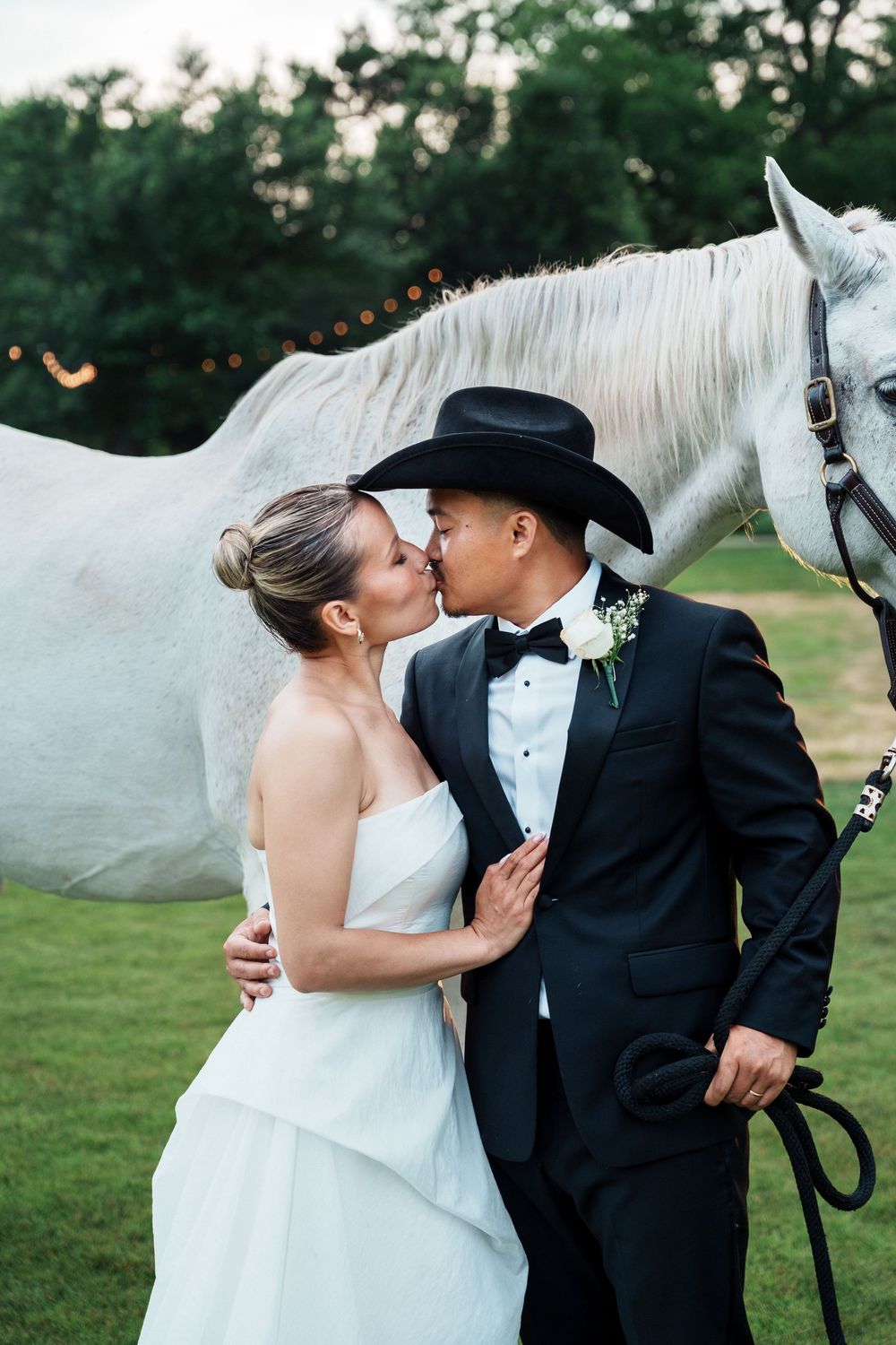 Bride and groom share a kiss outdoors in wedding attire, groom wearing black cowboy hat, white horse standing behind them.
