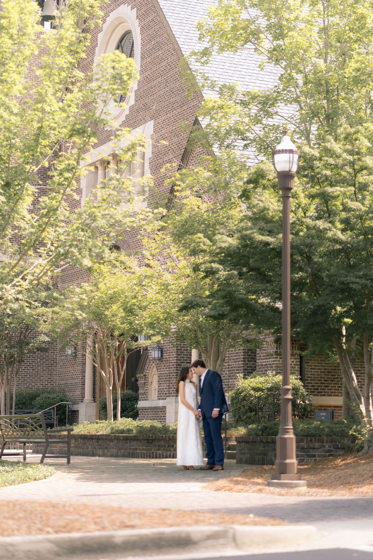 Couple sharing a romantic moment outside a brick church surrounded by lush green trees and a vintage street lamp.