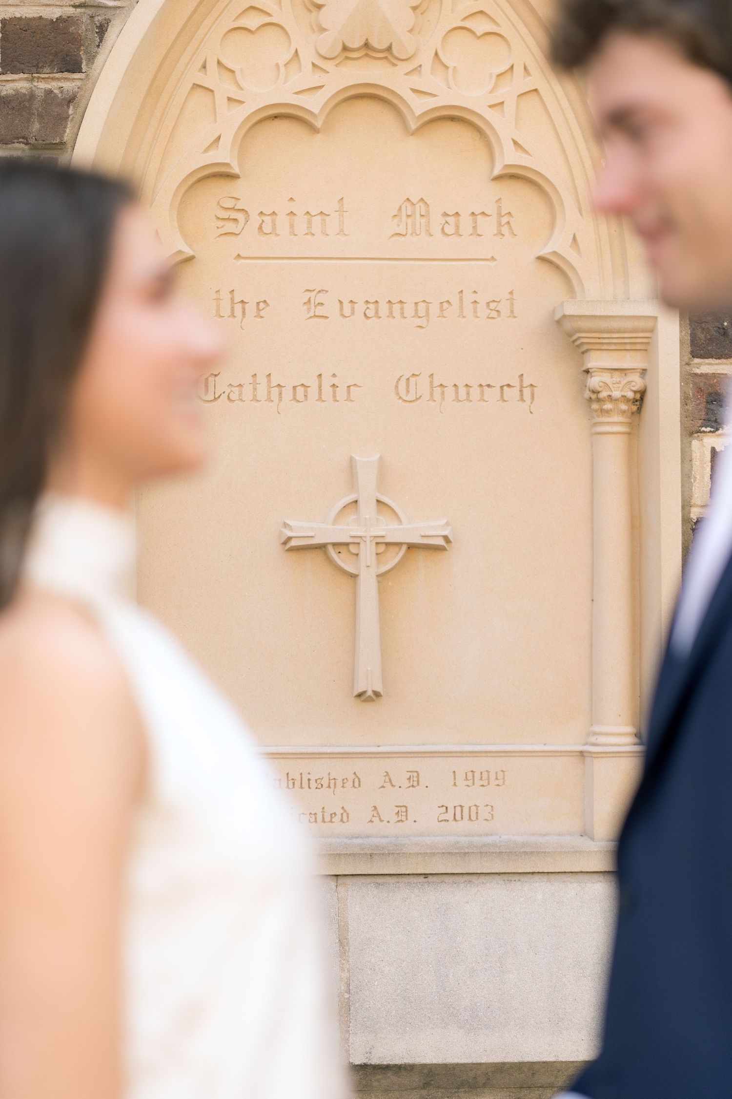 Couple posing in front of Saint Mark the Evangelist Catholic Church stone sign with cross.