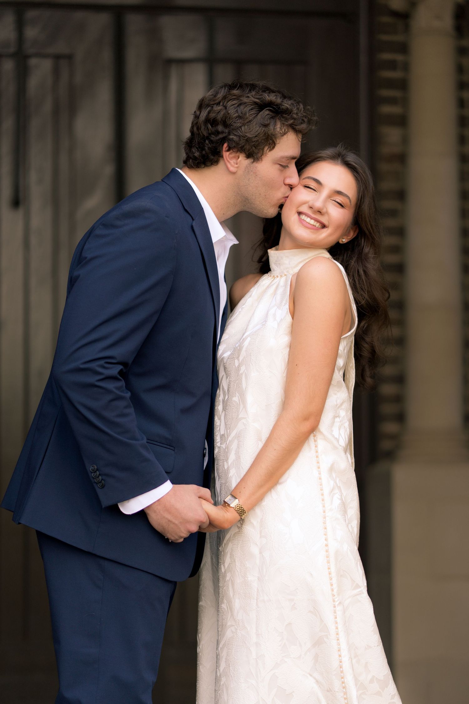 A groom in a navy suit kisses his smiling bride on the cheek as she wears a white lace dress.