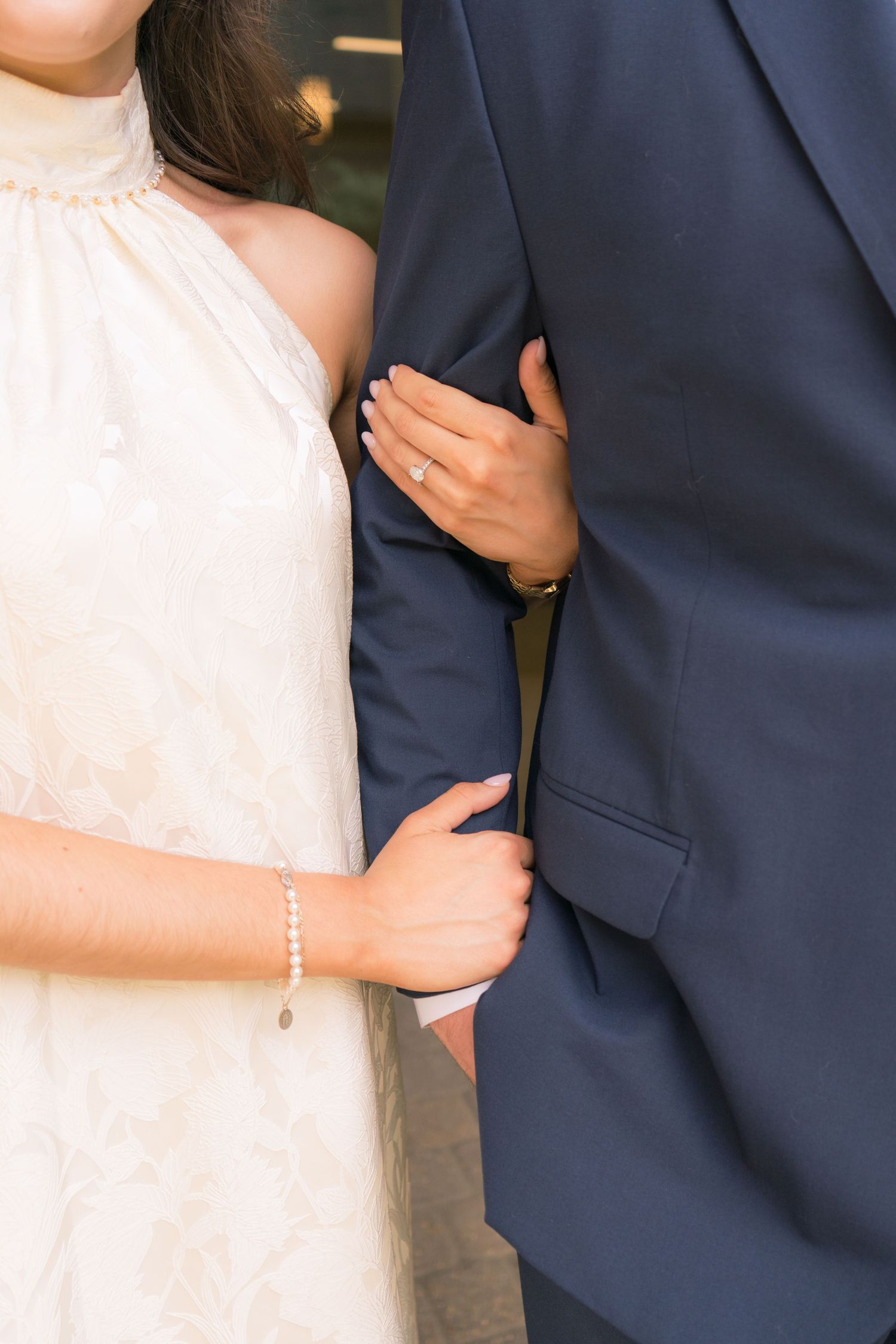 Close-up of bride in white halter dress with pearl bracelet holding groom's navy suit arm, wearing engagement ring.