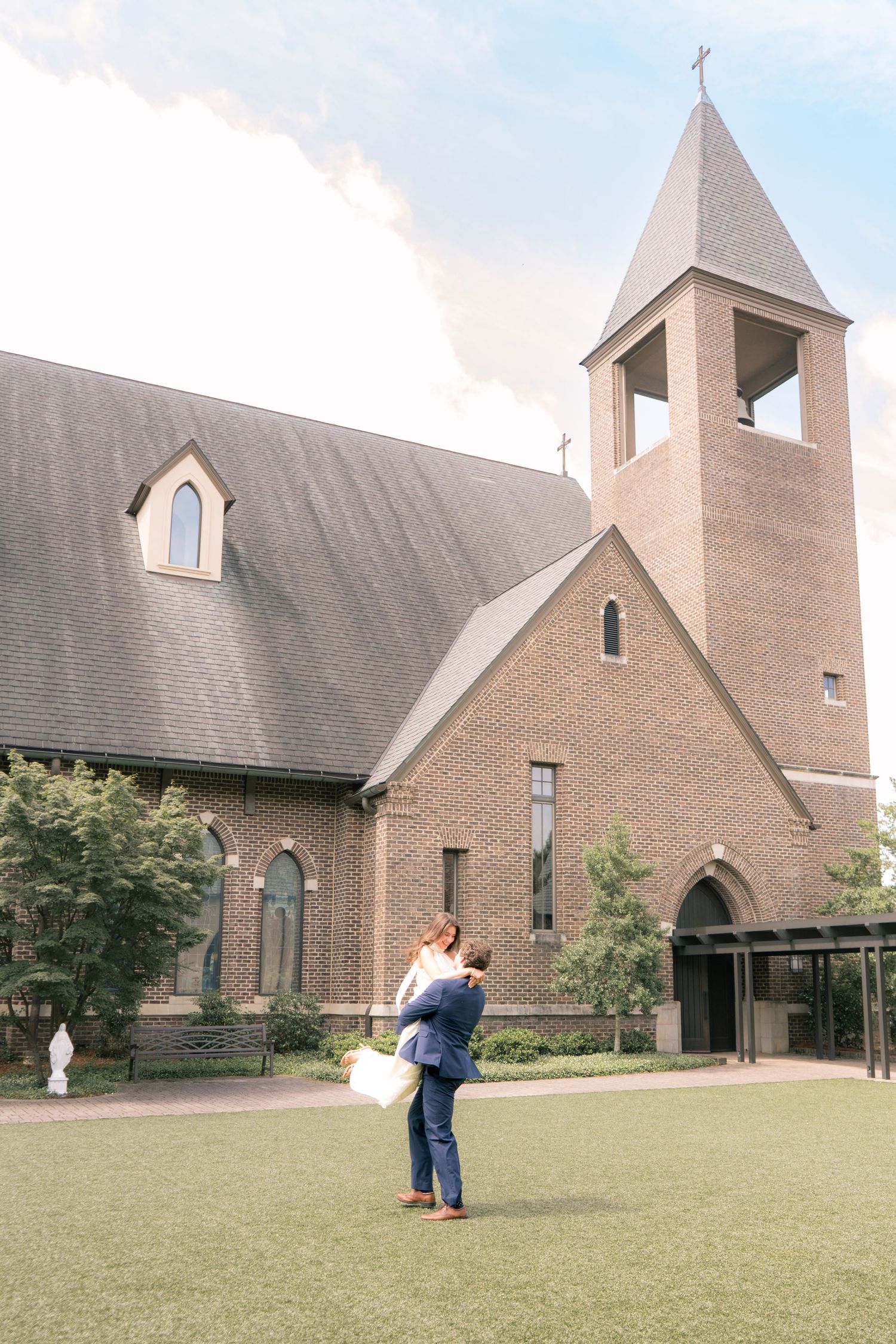 Couple embracing on green lawn outside a brick church with a bell tower and cross on a cloudy day.