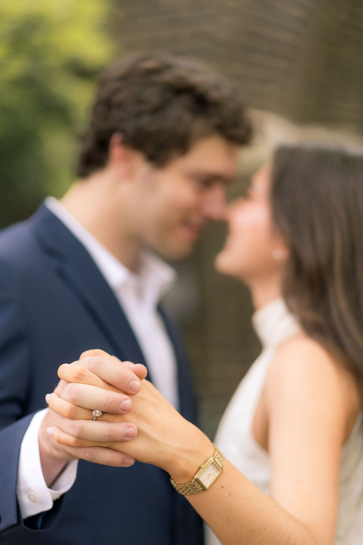 Couple holding hands with engagement ring and gold watch visible, sharing a romantic moment in soft focus outdoor setting.