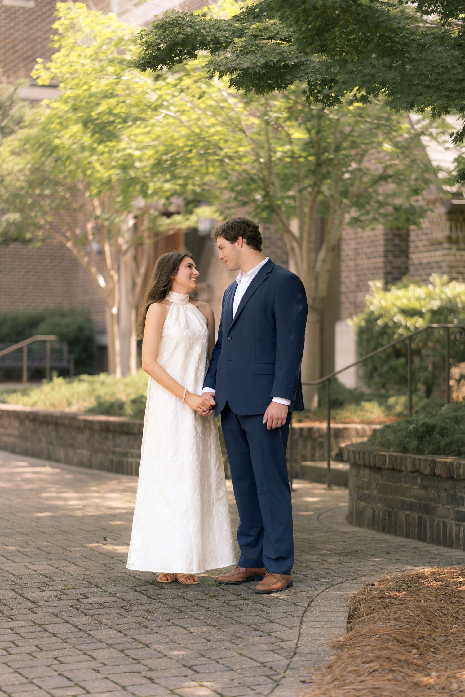 Couple in formal attire holding hands on a brick pathway surrounded by lush green trees and gardens.