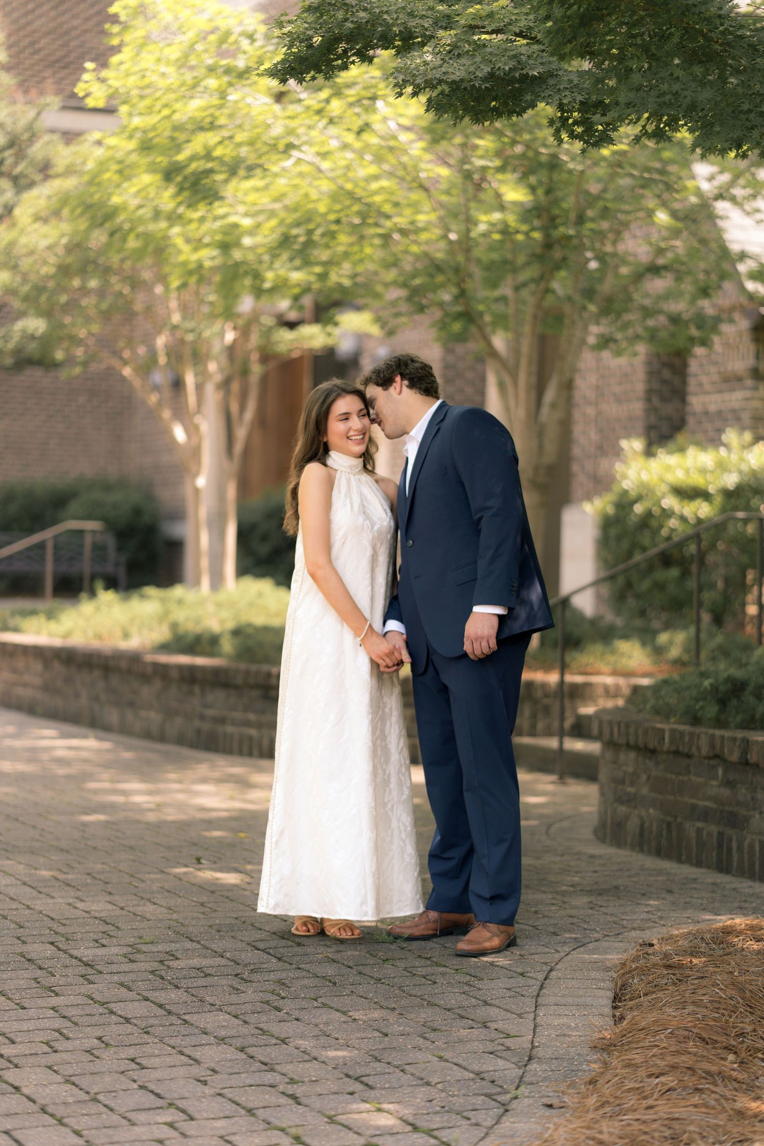Couple in formal wear holding hands on brick path, woman in white dress, man in navy suit, surrounded by trees.