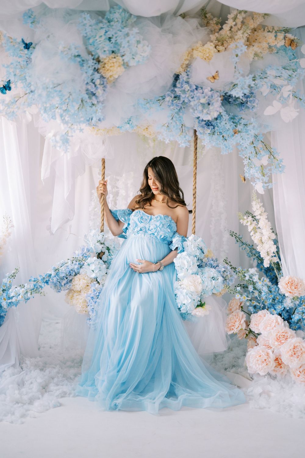 Ethereal maternity portrait with flowing light blue gown on swing surrounded by blue and peach floral arrangements.