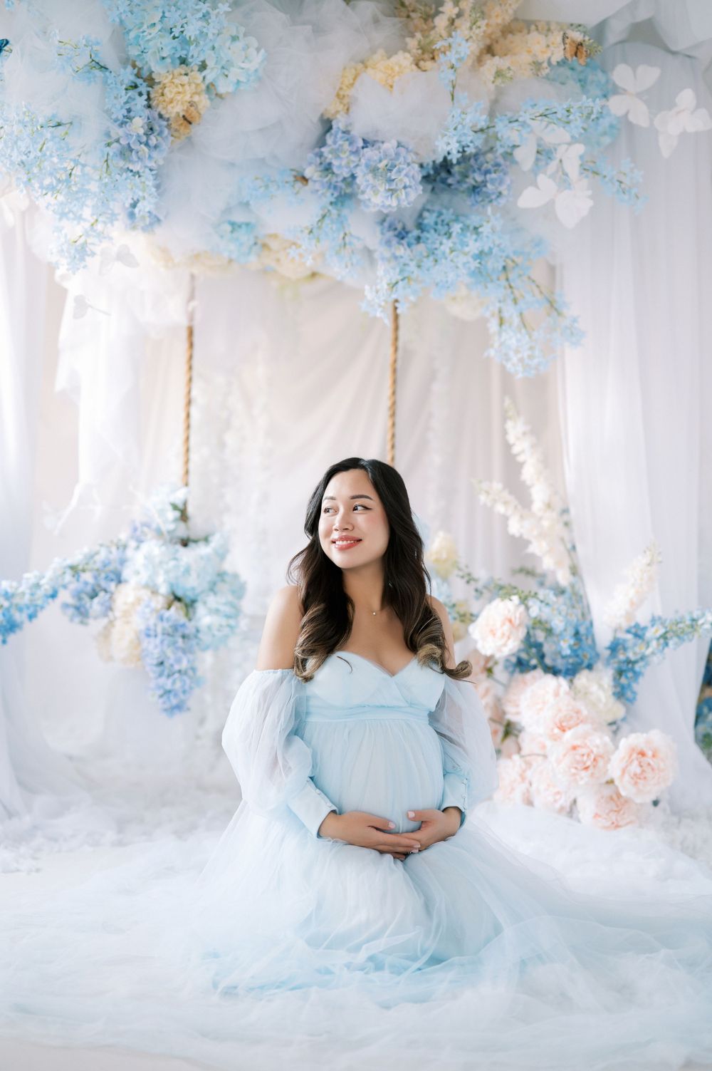 Ethereal maternity photo featuring soft blue florals and white drapery in a dreamy studio setting.