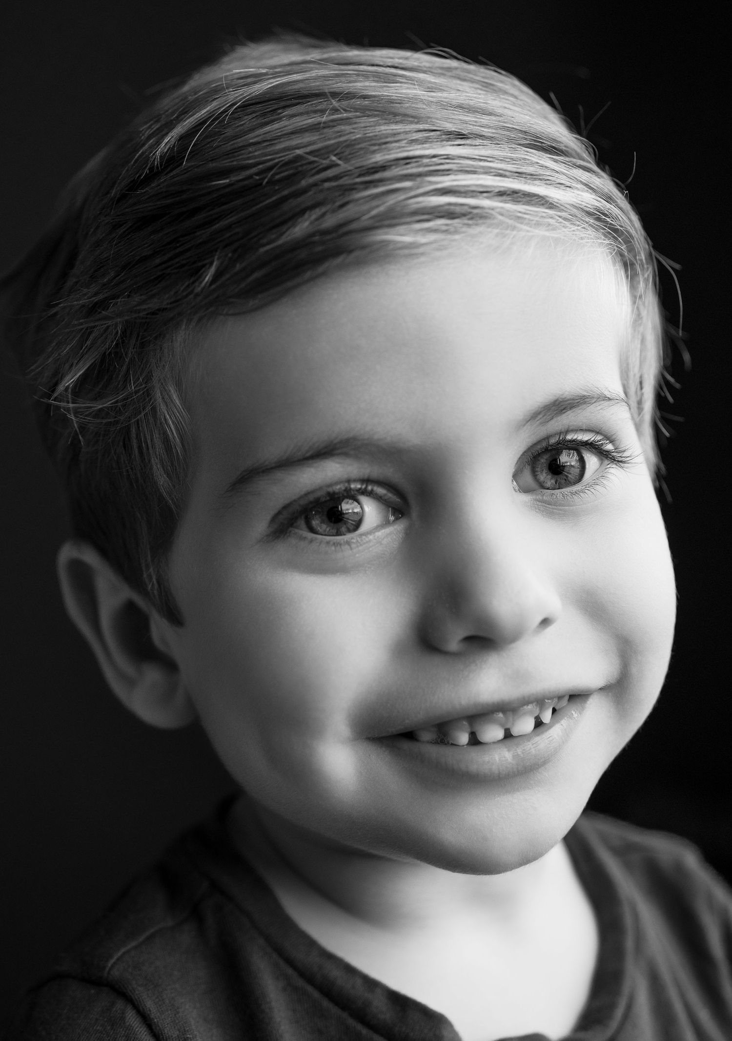 A black and white portrait of a smiling young child with a bright expression and short hair against a dark background.