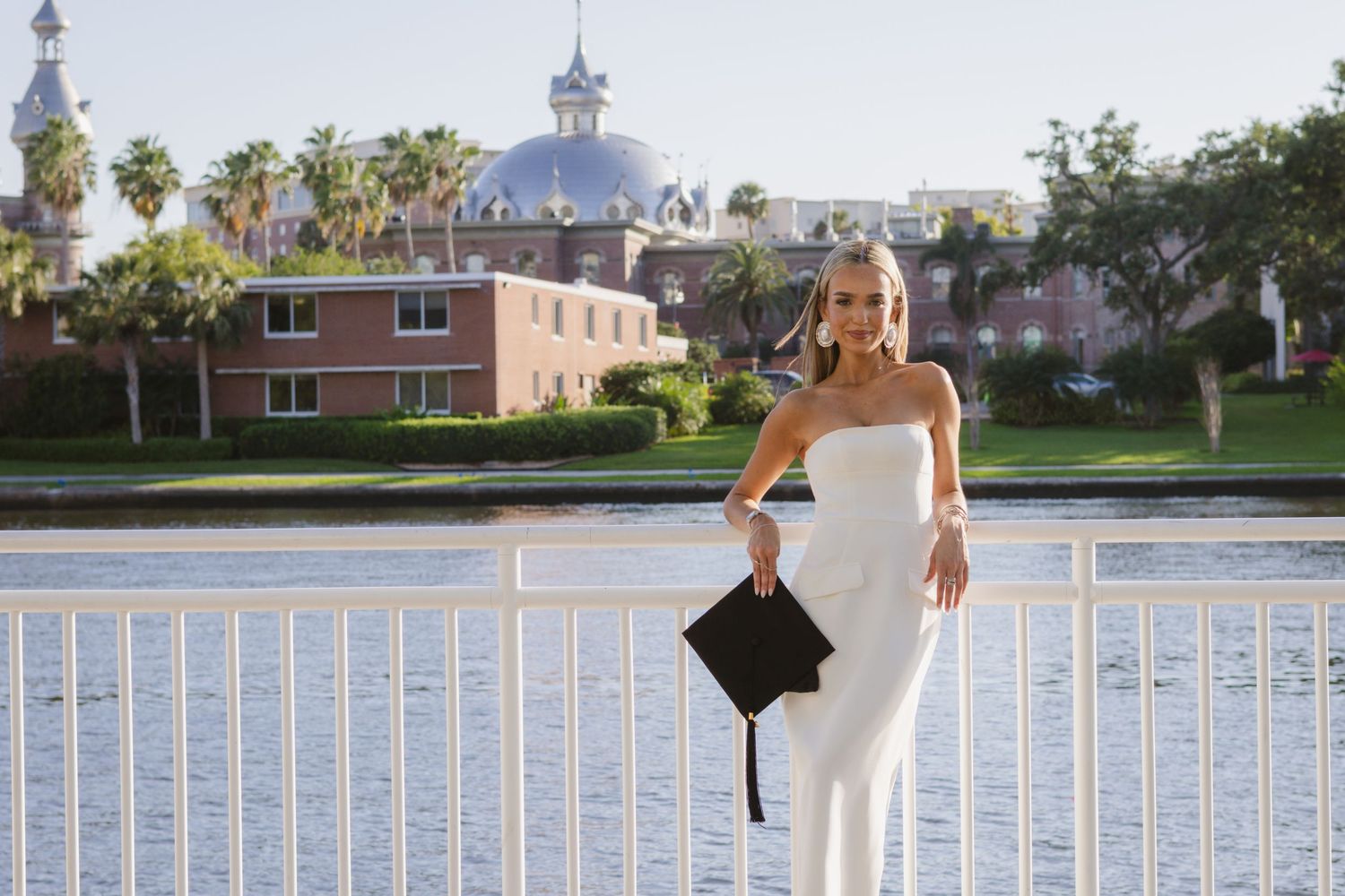 Leaned on the white railing at Curtis Hixon Park, a woman with her graduation cap poses. The Tampa campus is seen over the water behind her.