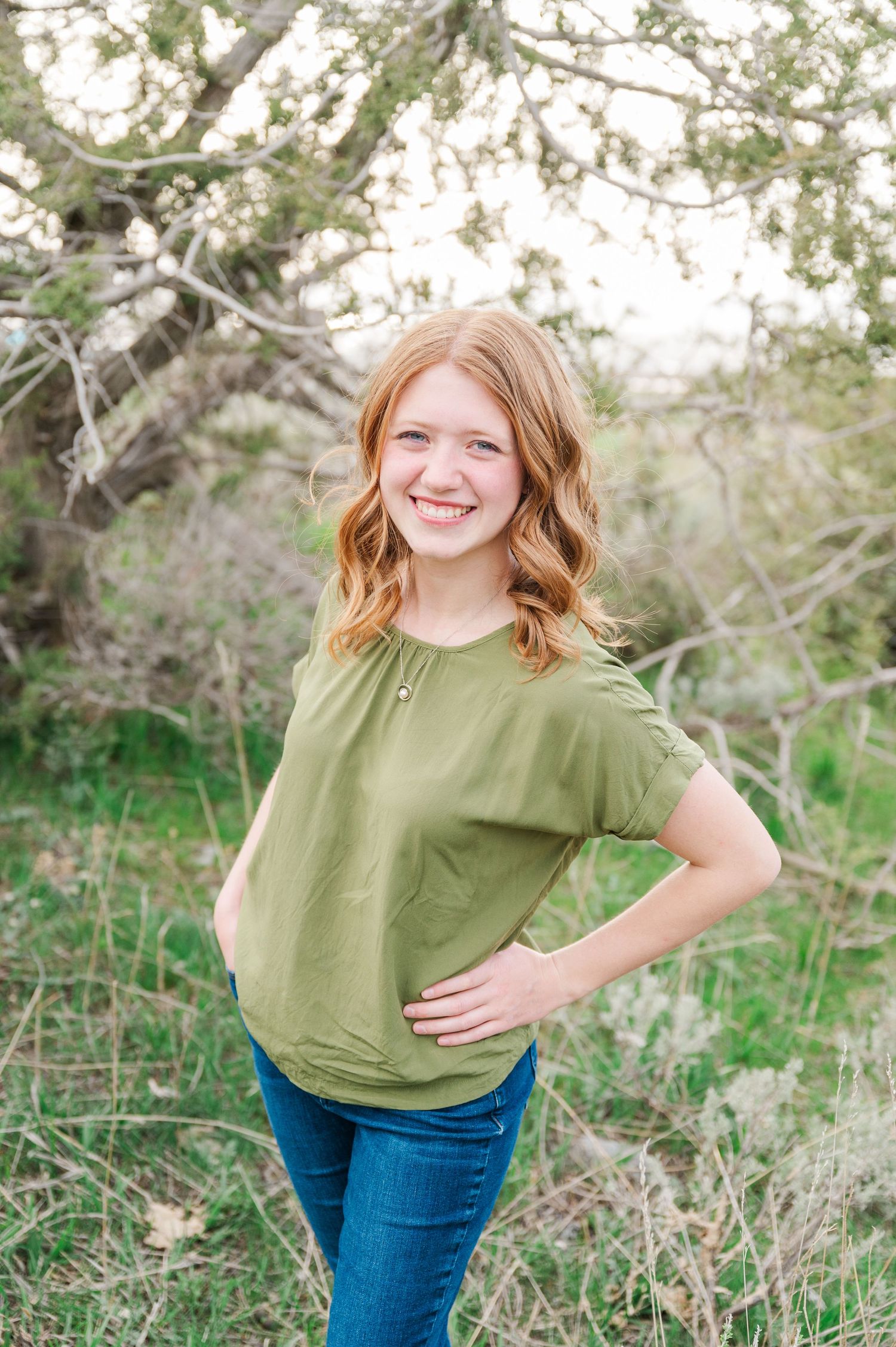 Casual outdoor portrait of someone in an olive green top and jeans against a natural background.