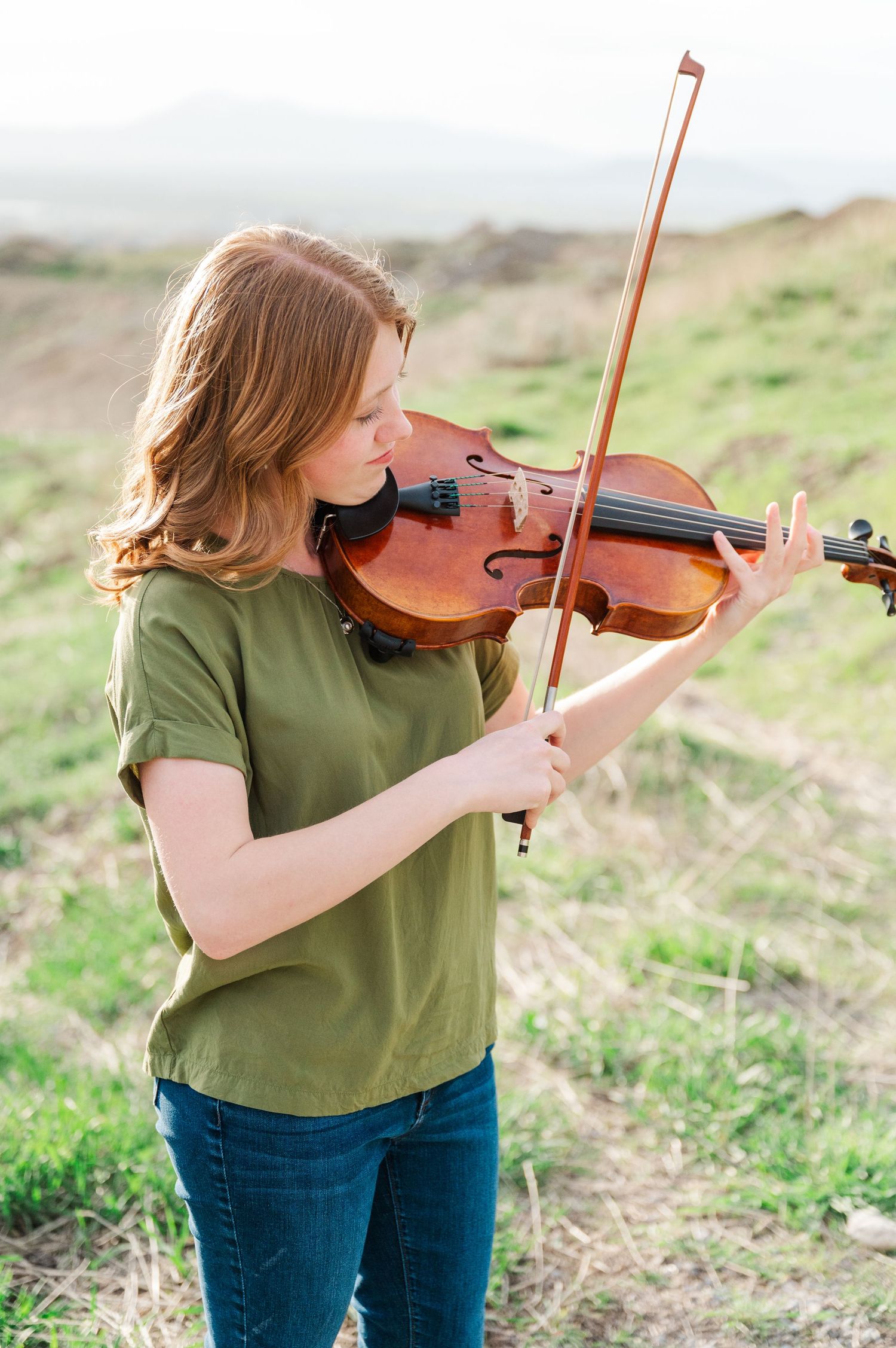 Musician playing violin outdoors on a hillside wearing casual green top and jeans.