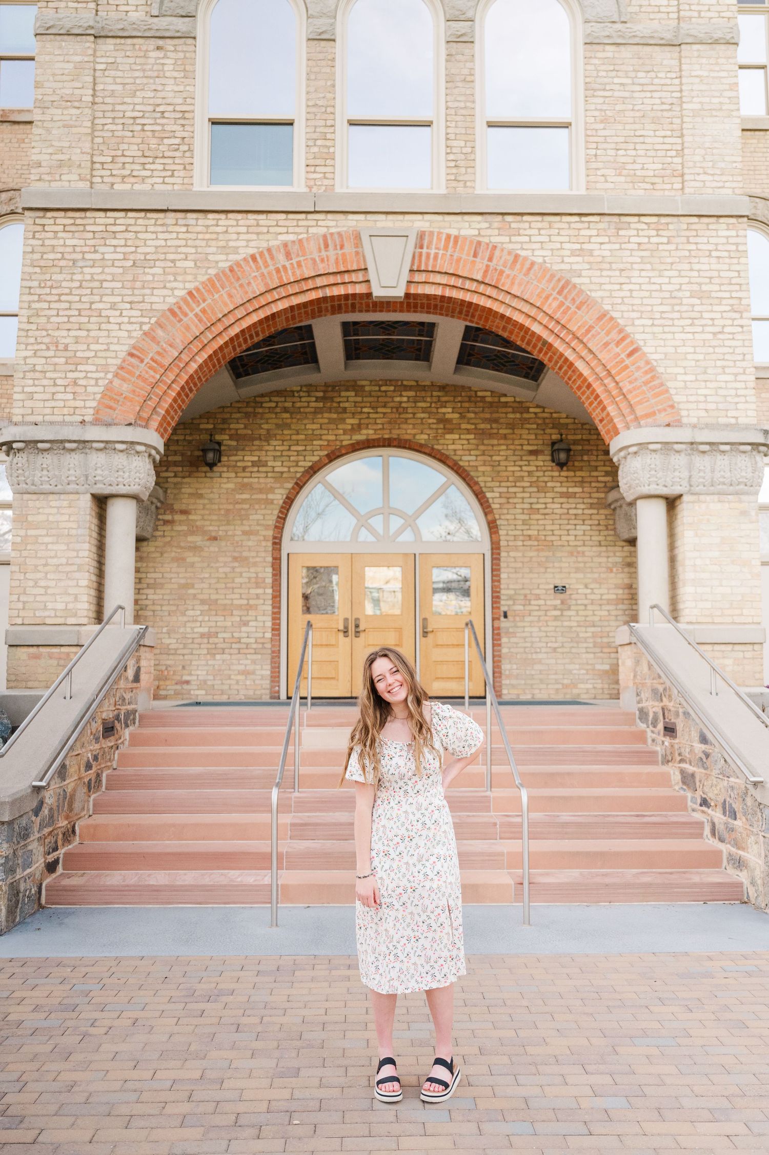 A graduate in a white polka dot dress poses on steps in front of a historic brick building with arched doorways.