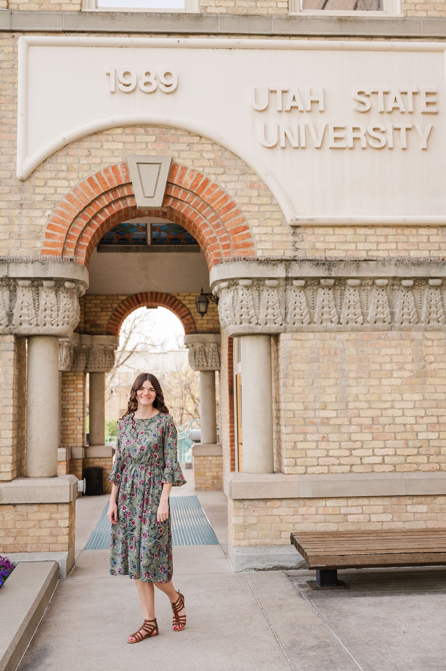 Graduate poses under Utah State University archway in floral dress.