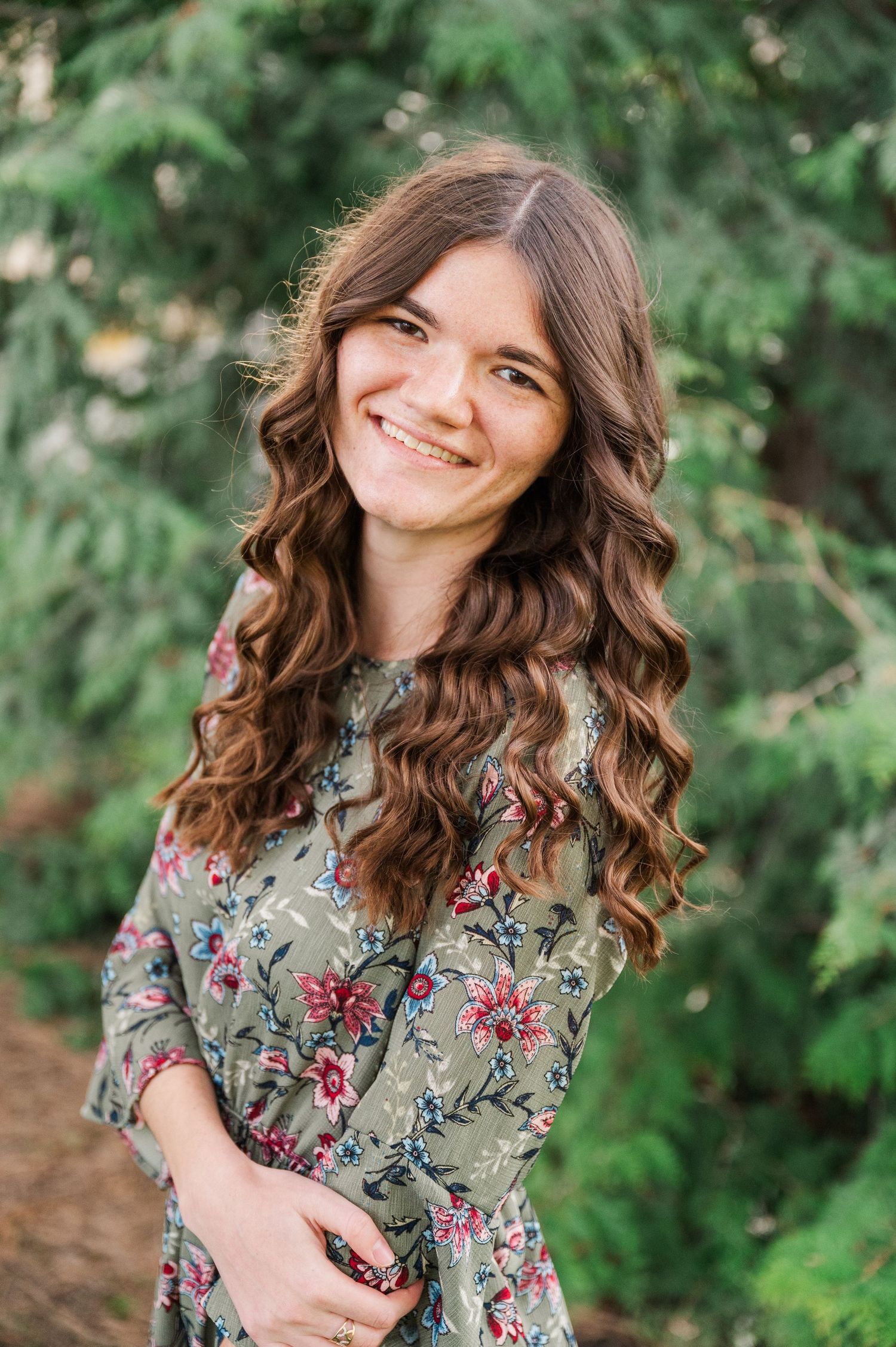 Close up portrait outdoors featuring wavy hair and a floral print dress against evergreen trees.