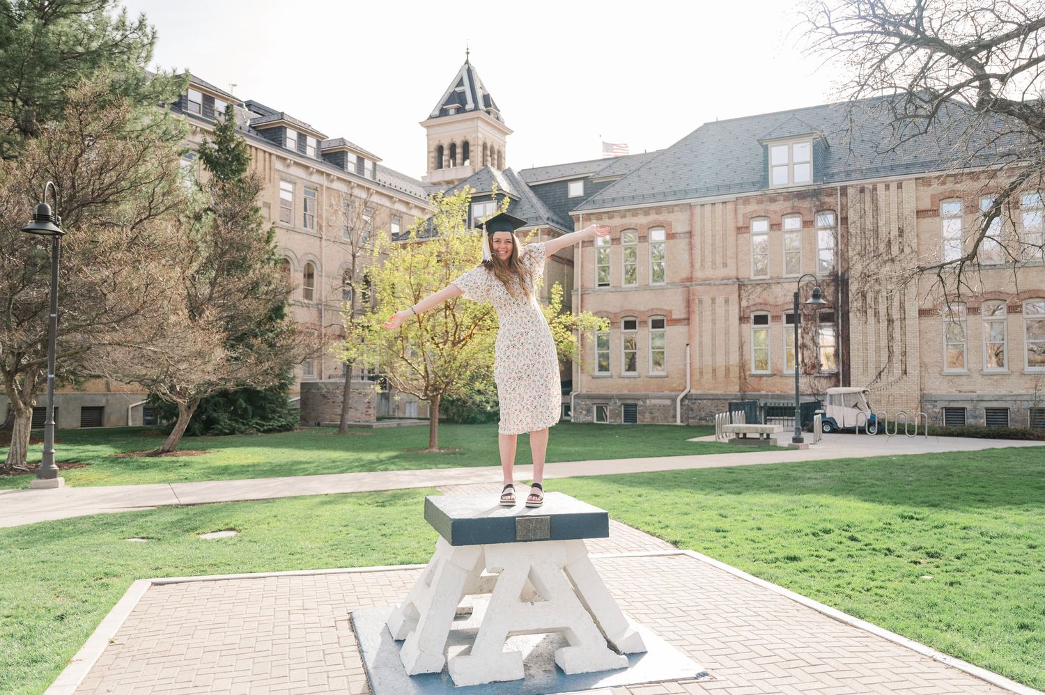 Graduate in white dress standing on a blue and white platform with historic university buildings in background.