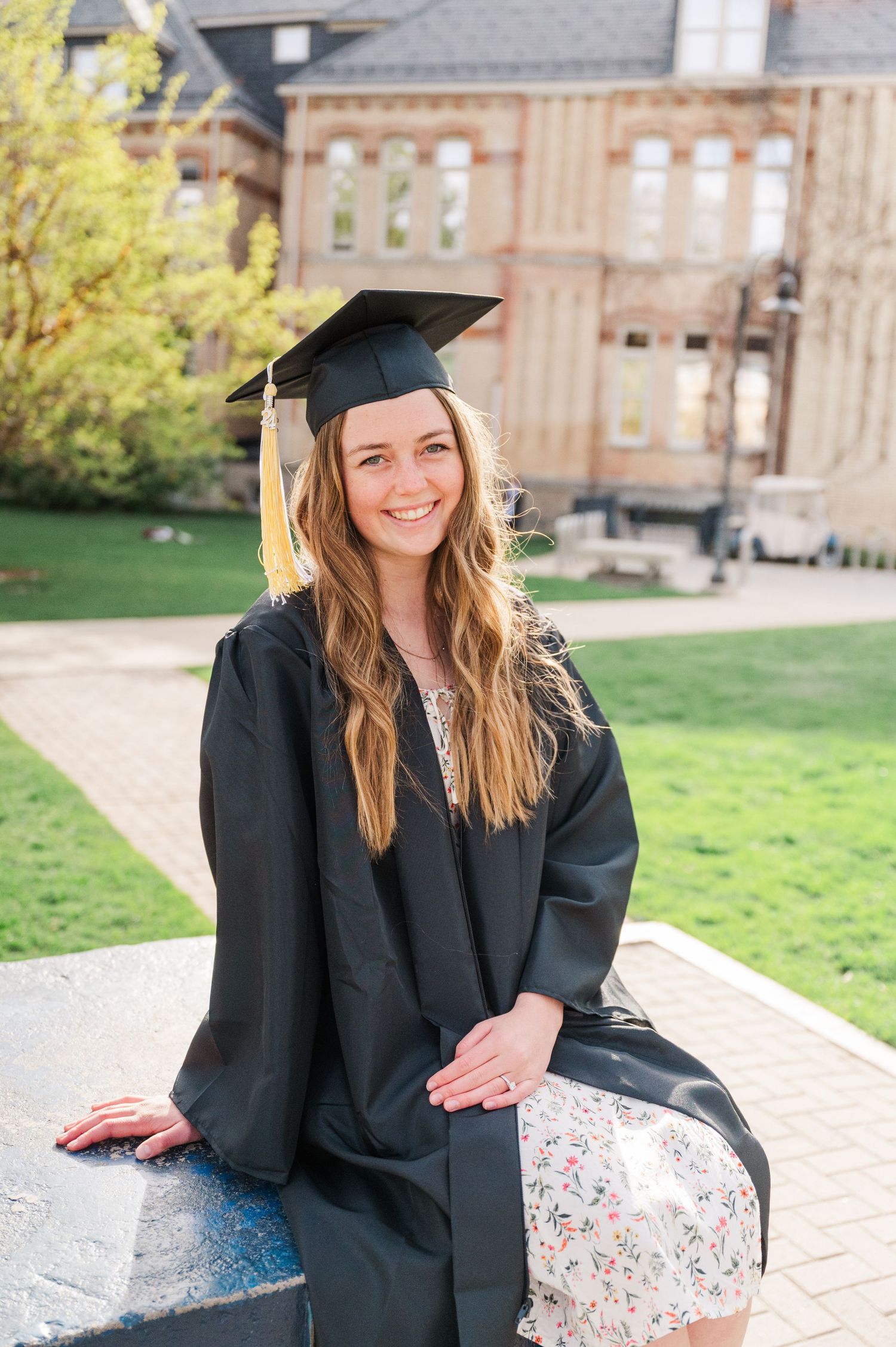 Graduate in cap and gown sits on outdoor campus furniture during commencement.