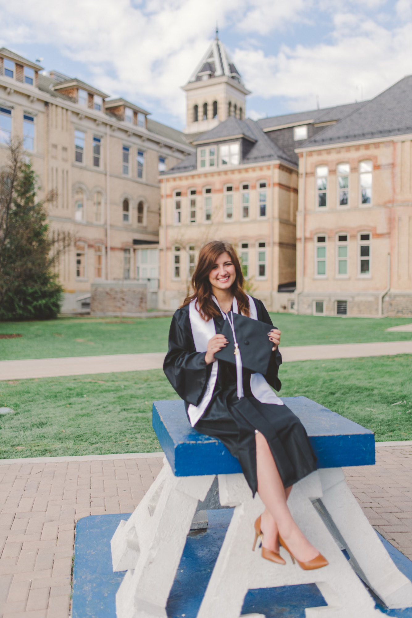 Graduate in academic regalia sits on blue and white bench with historic college architecture behind.