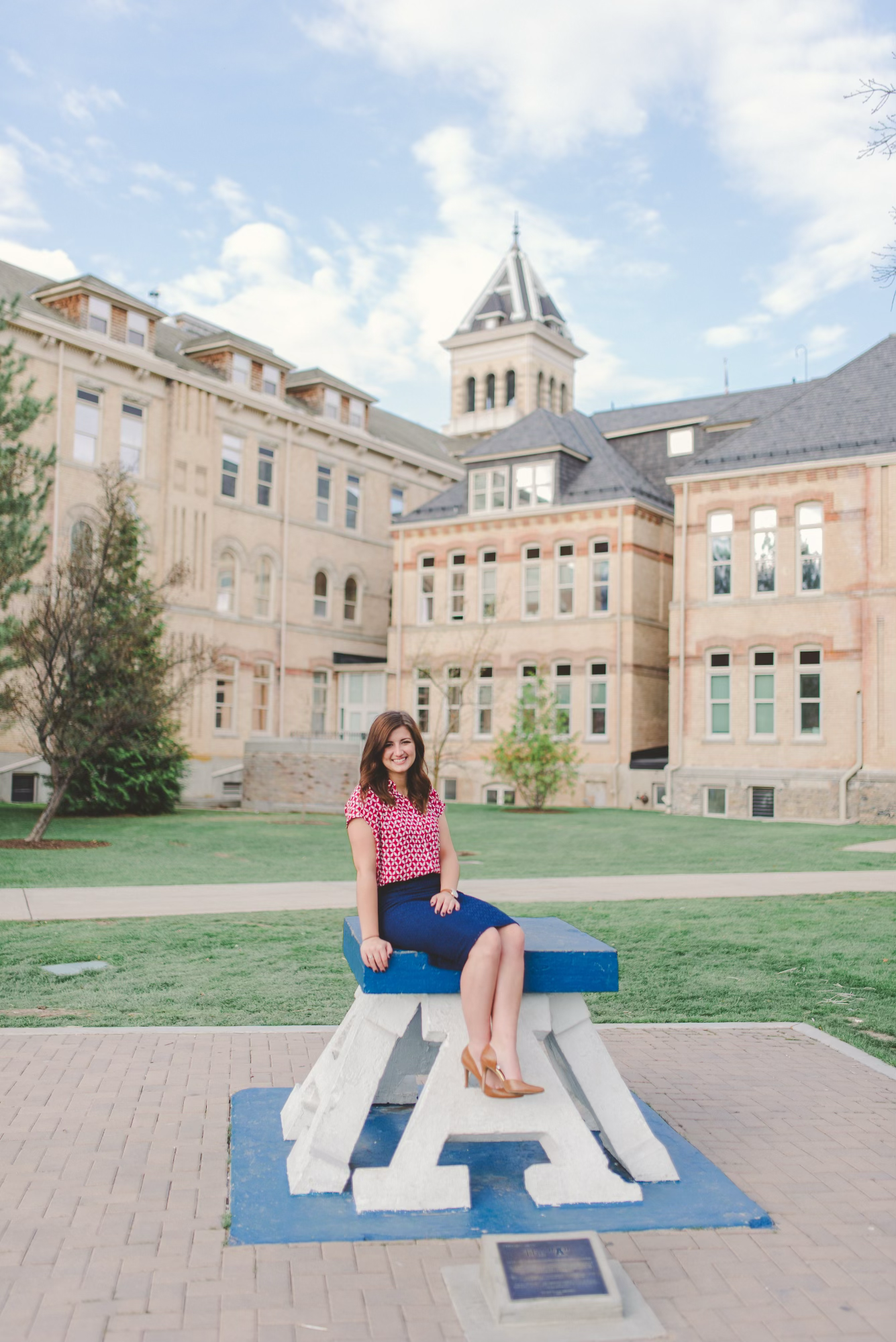 Graduate in pink top and blue skirt sits on campus bench with historic university tower in background.