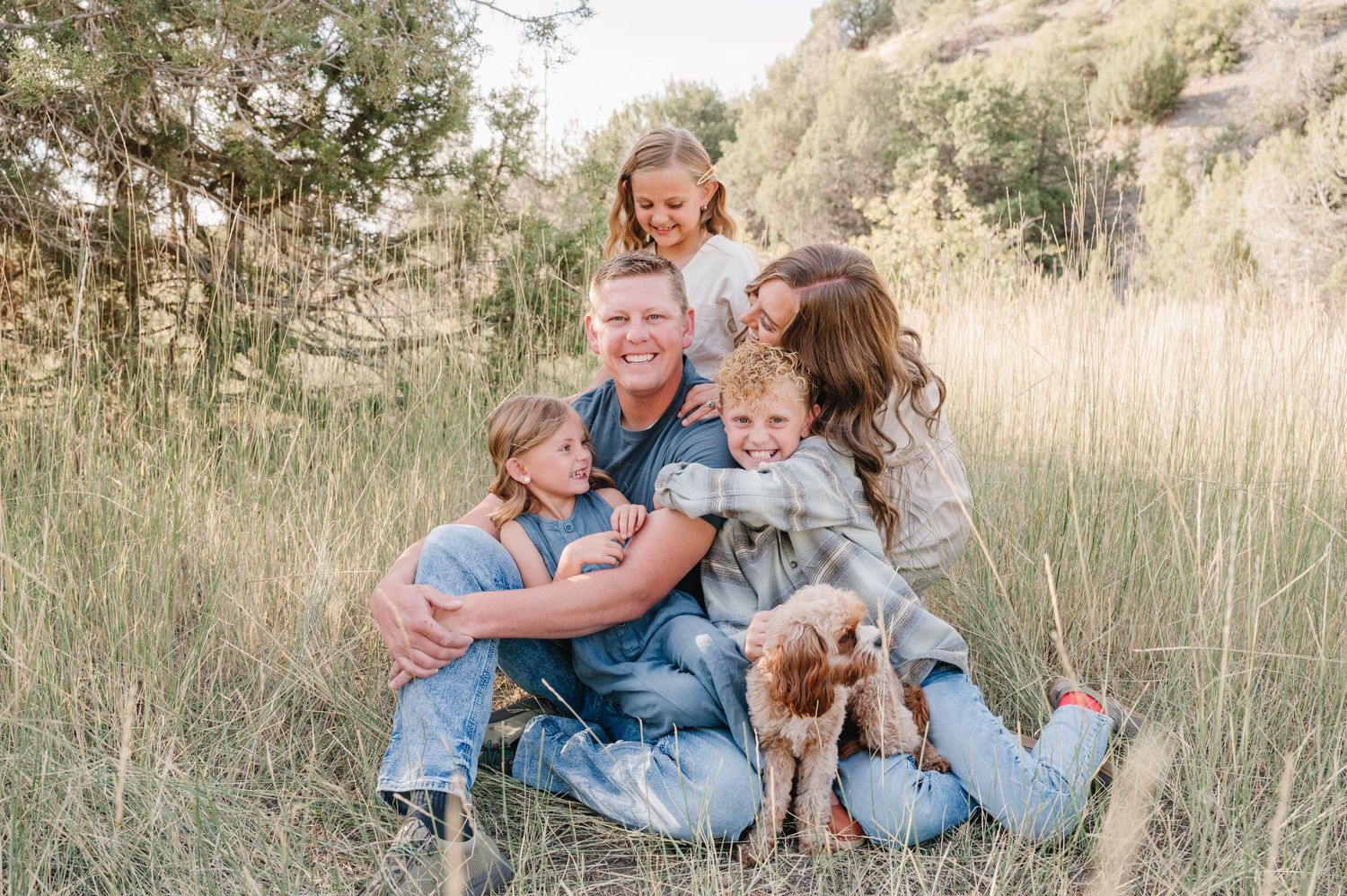 Family cuddles together with their dog in golden mountain meadow setting.