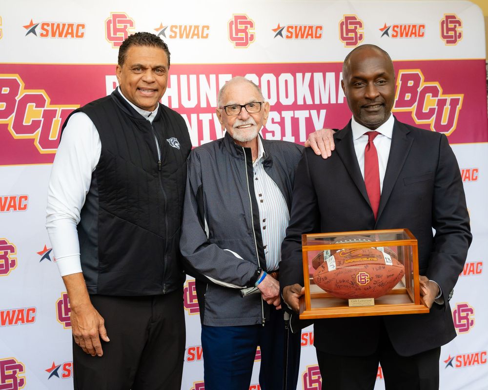 Three people pose with commemorative football plaque at university branded press backdrop.