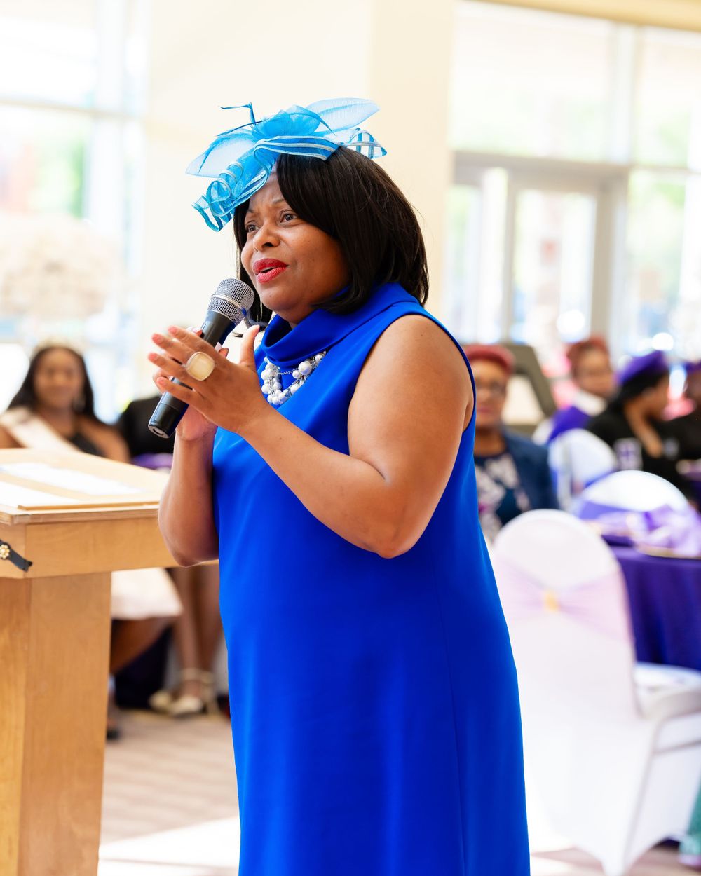 Speaker in blue dress and light blue fascinator addresses audience from wooden podium.