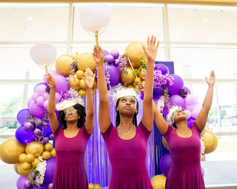 Three performers in magenta dresses raising hands under purple and gold balloon arch.