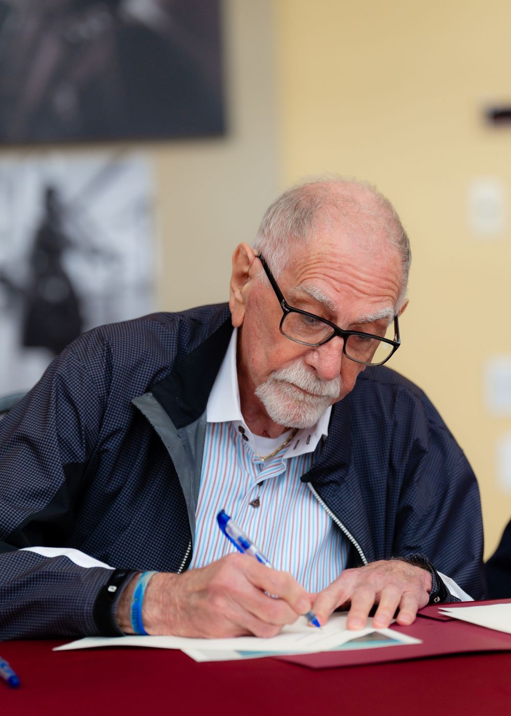 Senior person in navy jacket signs document while seated at desk.