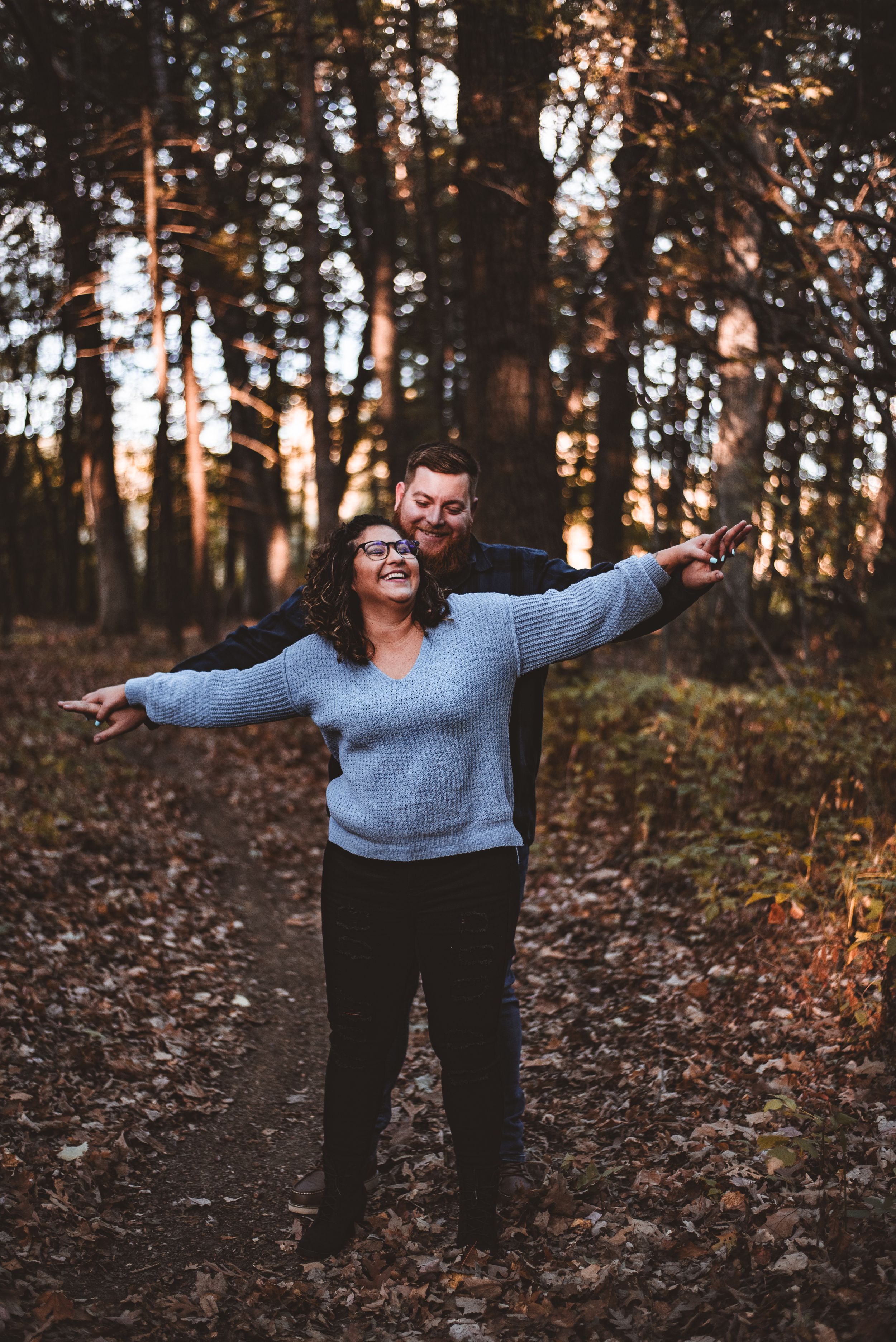 Playful couple pose with outstretched arms in a fall forest setting.