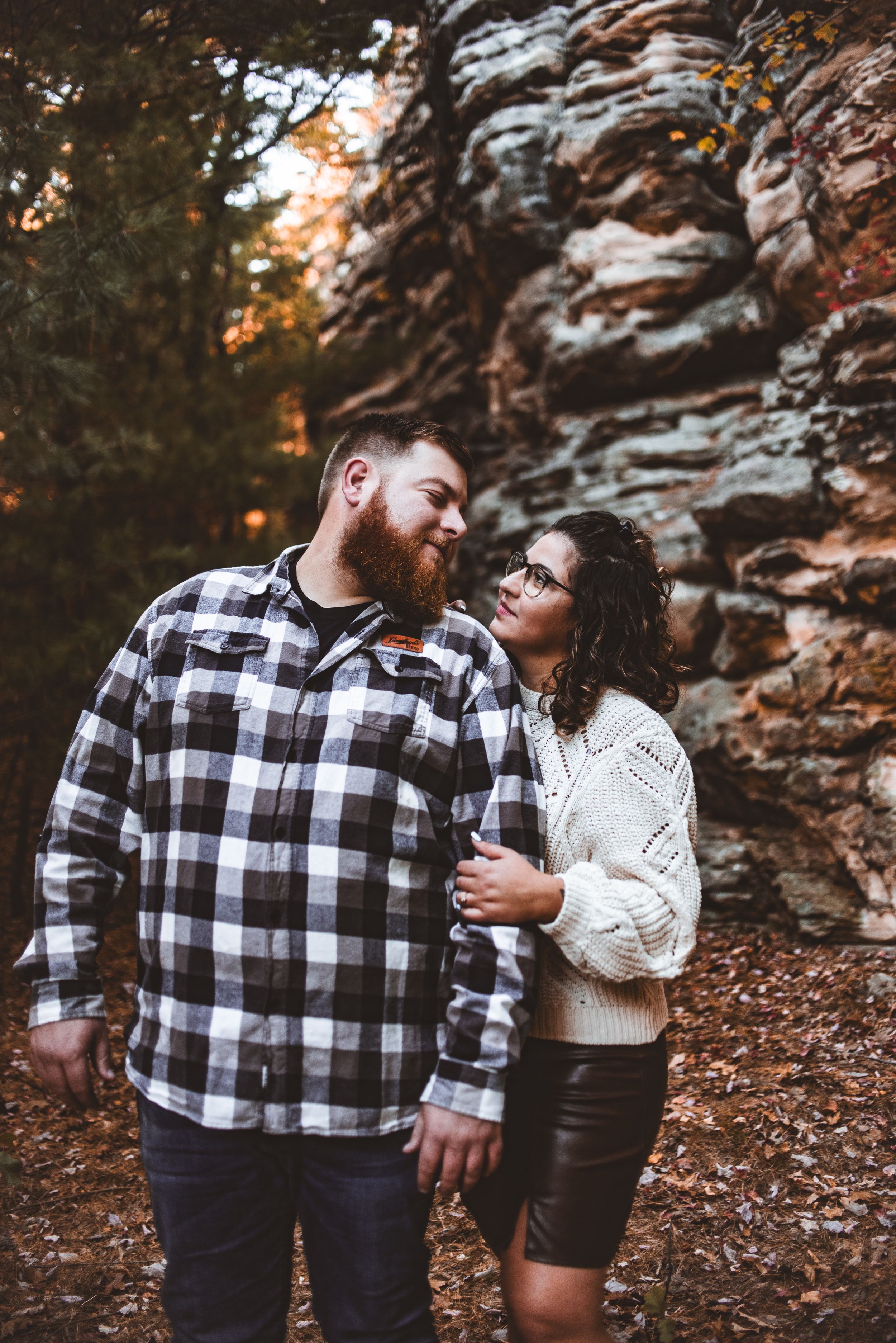 Couple in plaid and knit sweaters pose intimately against autumn rock formation backdrop.