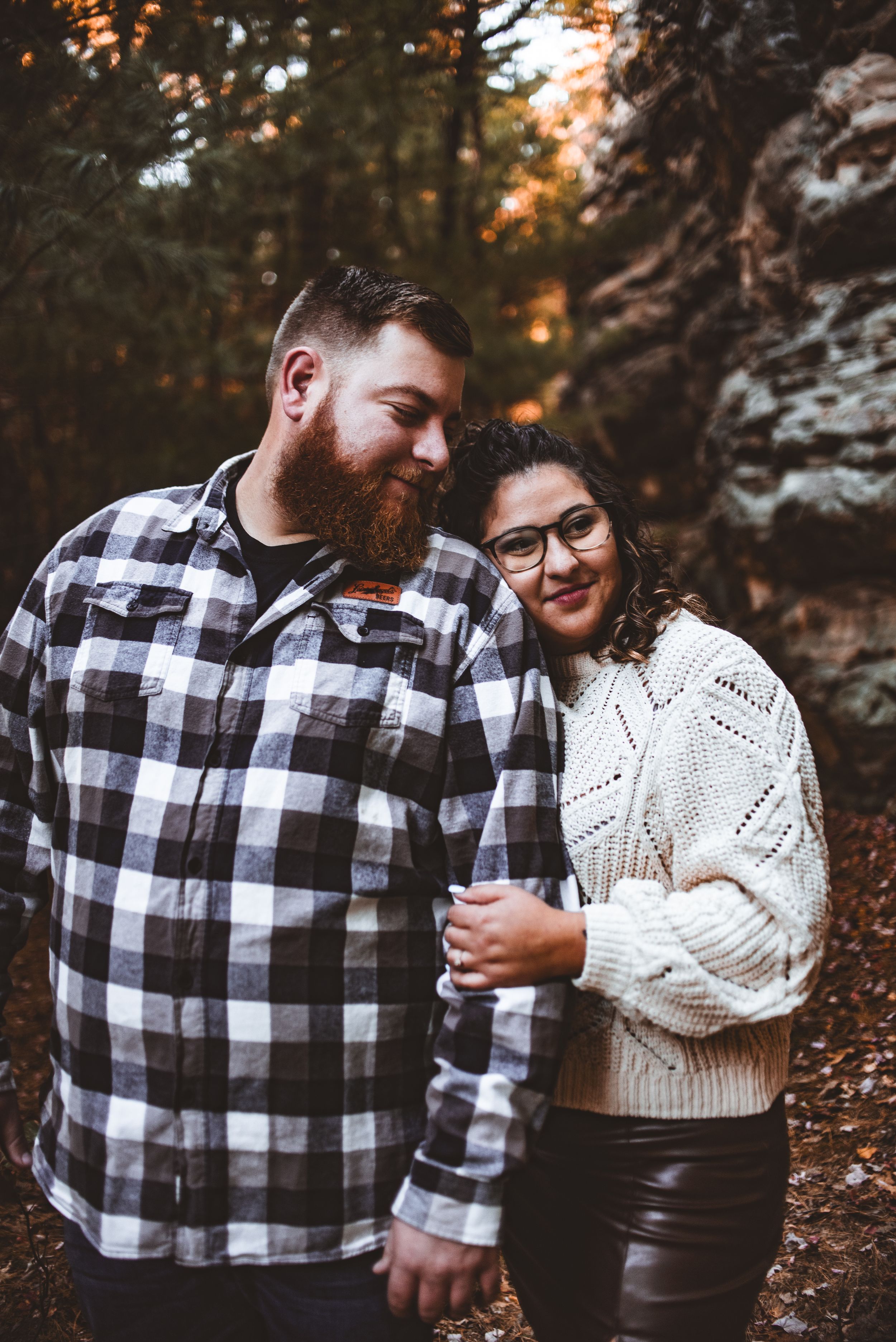 A couple poses for a photo in the forest wearing contrasting plaid and knit sweater outfits in autumn.