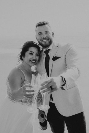 A happy couple in wedding attire toasts with champagne glasses in a black and white photo.