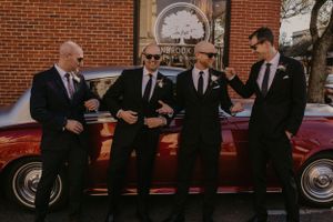 Four individuals in formal black suits stand together near a vintage red car against a brick building background.