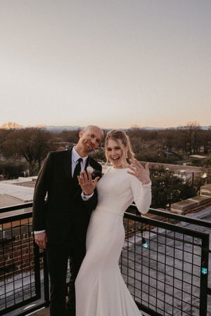 A couple celebrates on a rooftop balcony at sunset, with the bride wearing a fitted white gown and groom in dark suit.