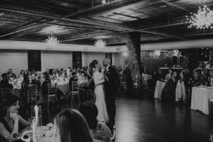 A black and white photograph shows a couple dancing at an elegant indoor reception with tables and guests around them.