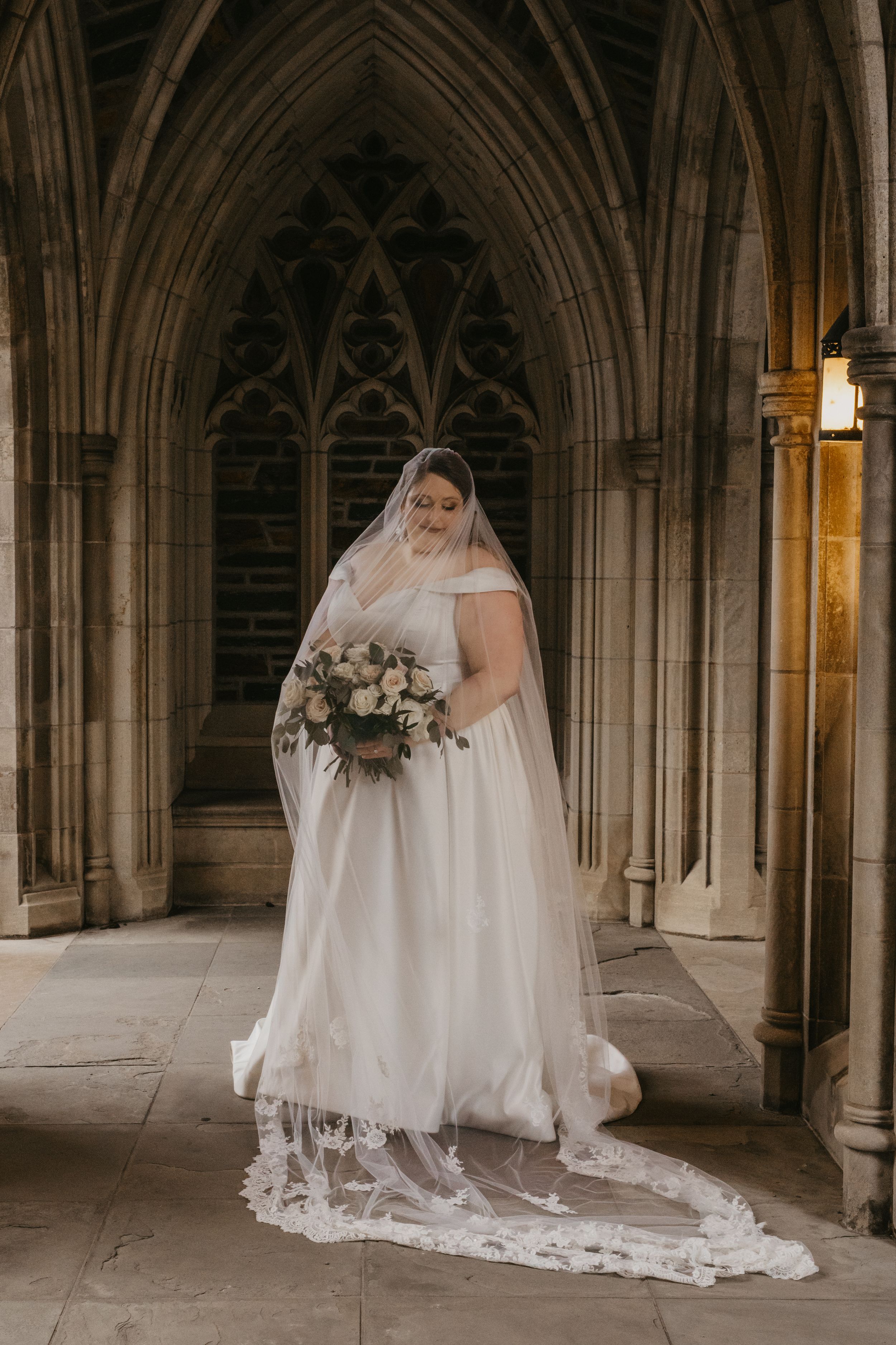A bride in a flowing white gown and long lace veil poses with a pink bouquet beneath gothic stone arches.