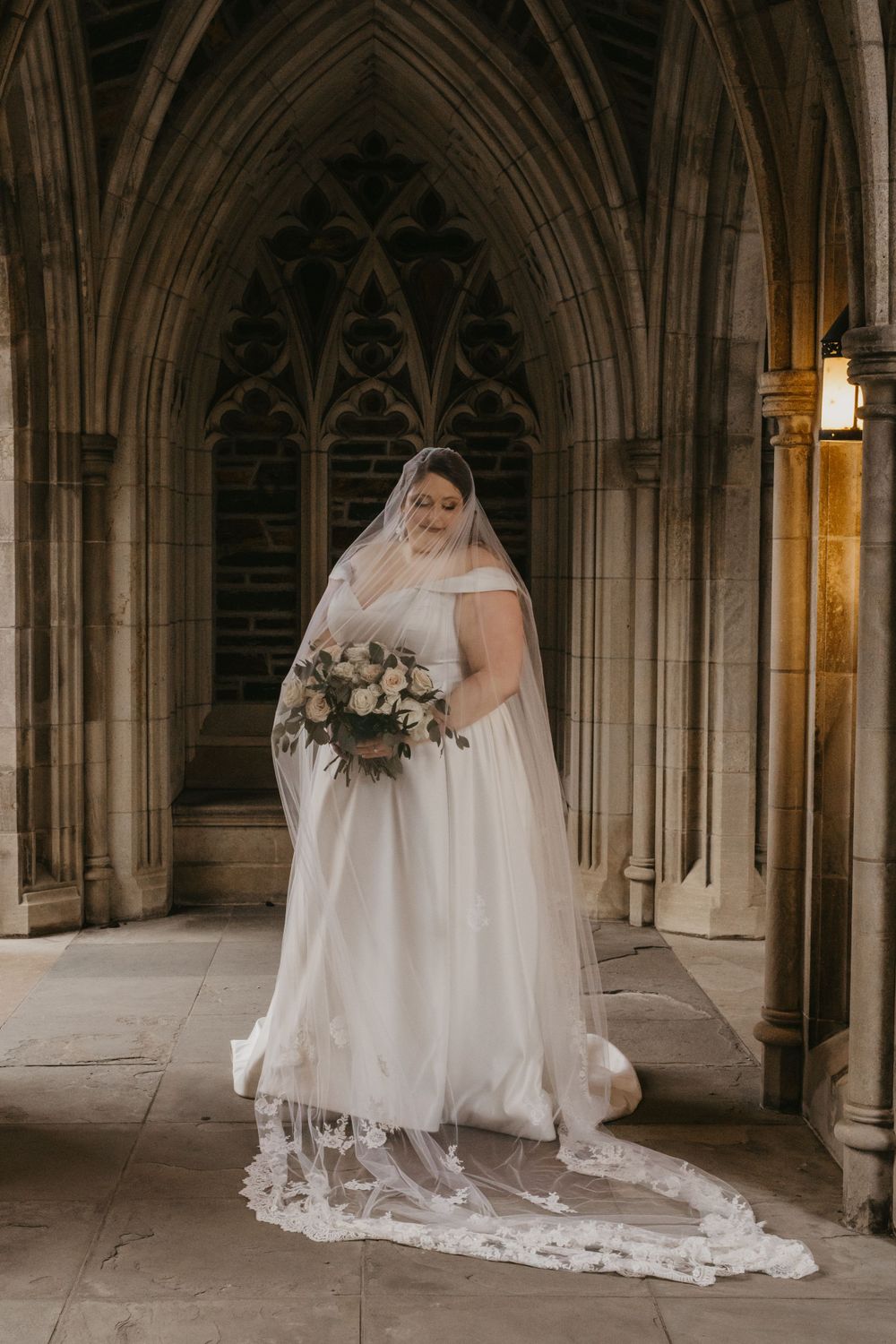 A bride in a flowing white gown and long lace veil poses with a pink bouquet beneath gothic stone arches.