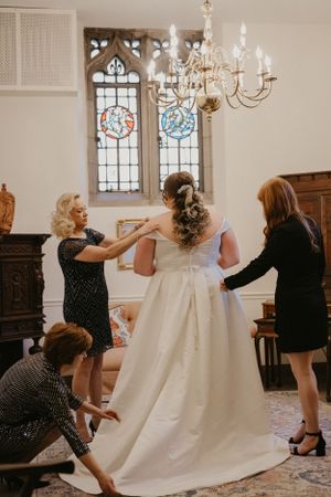 Wedding party helps with final dress adjustments in elegant chapel room with stained glass windows and chandelier.