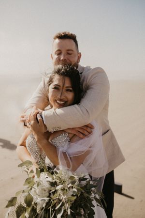 A joyful couple embraces on their wedding day at the beach, with the bride holding a white and green floral bouquet.