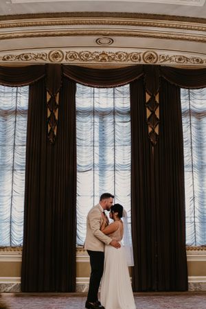 Romantic wedding couple stands before ornate curtains with elegant gold-trimmed crown molding in a grand ballroom.