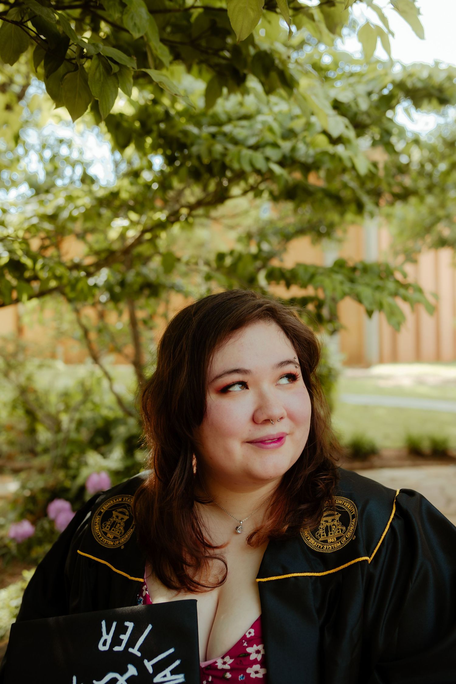 Graduate wearing black academic regalia with gold trim poses outdoors on sunny day.