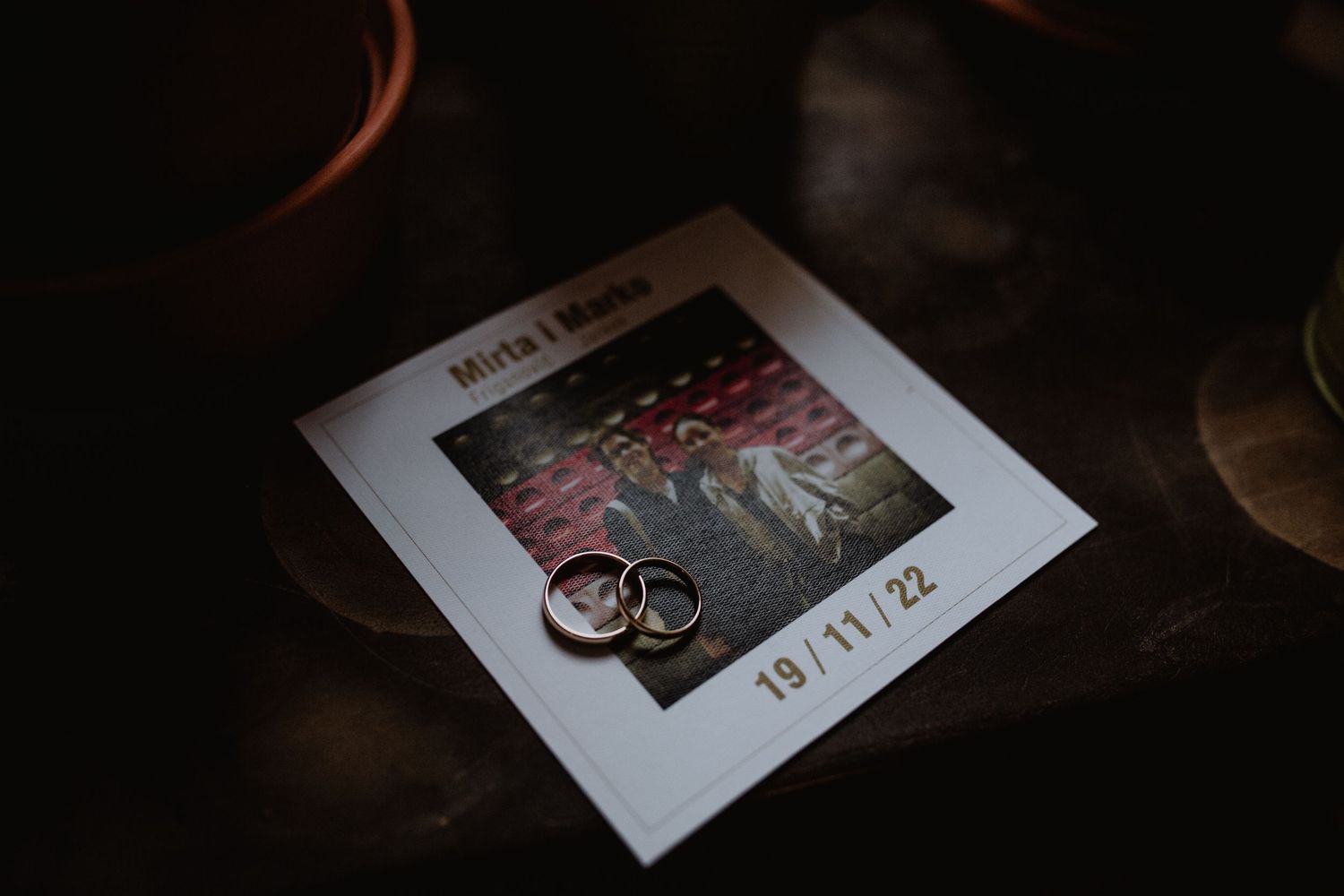 A close-up of wedding rings beside a polaroid photo in dramatic low lighting.