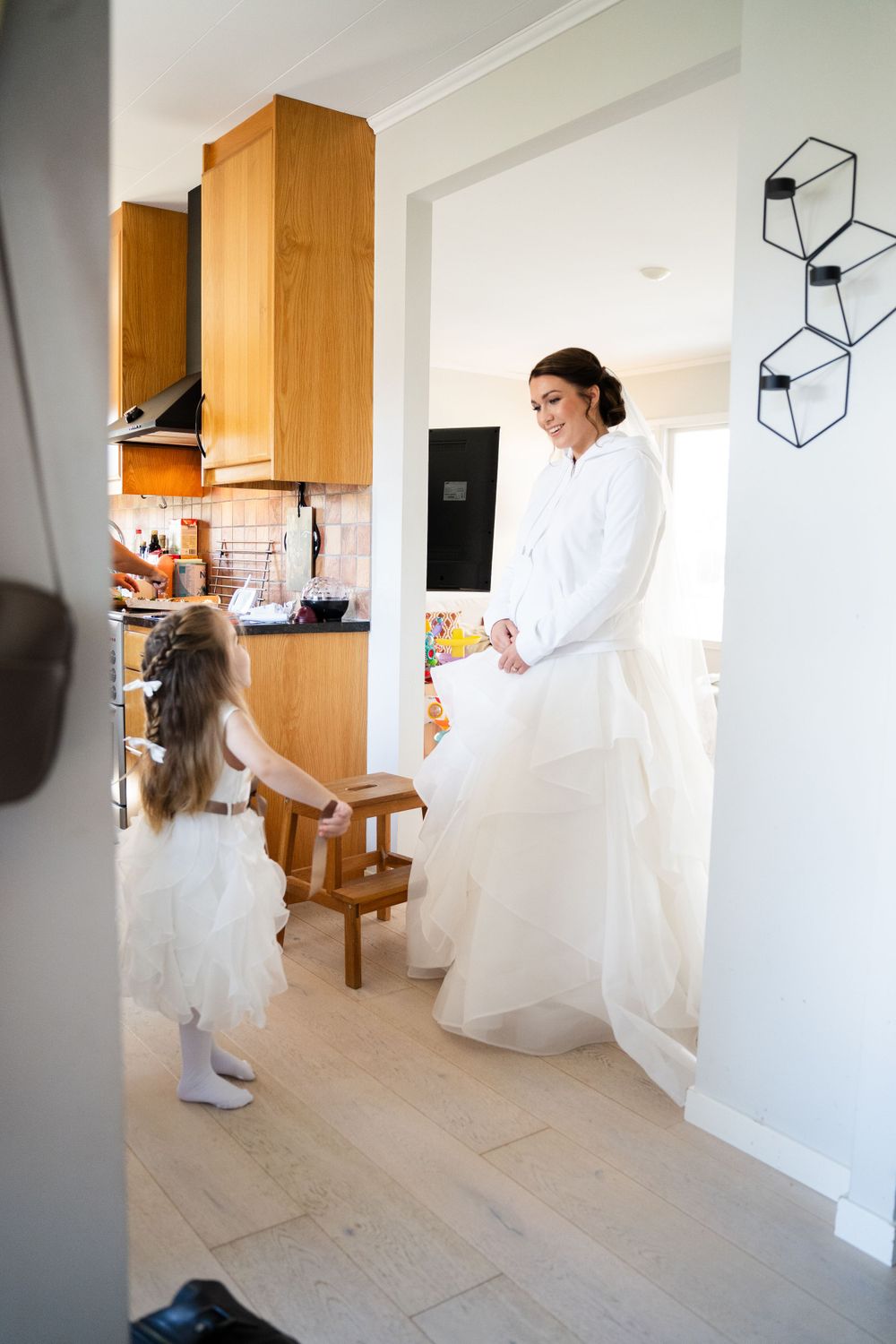 A bride in white gown interacts with young flower girl in modern kitchen setting.