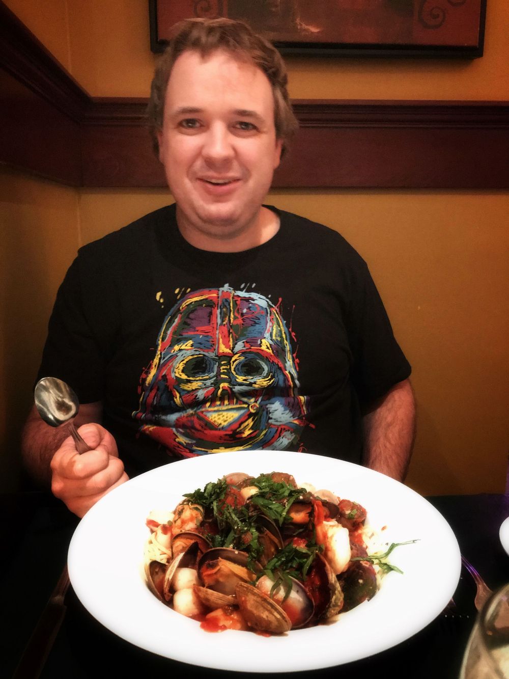 A Star Wars fan with a Darth Vader t-shirt sits at a restaurant with a gourmet meal.
